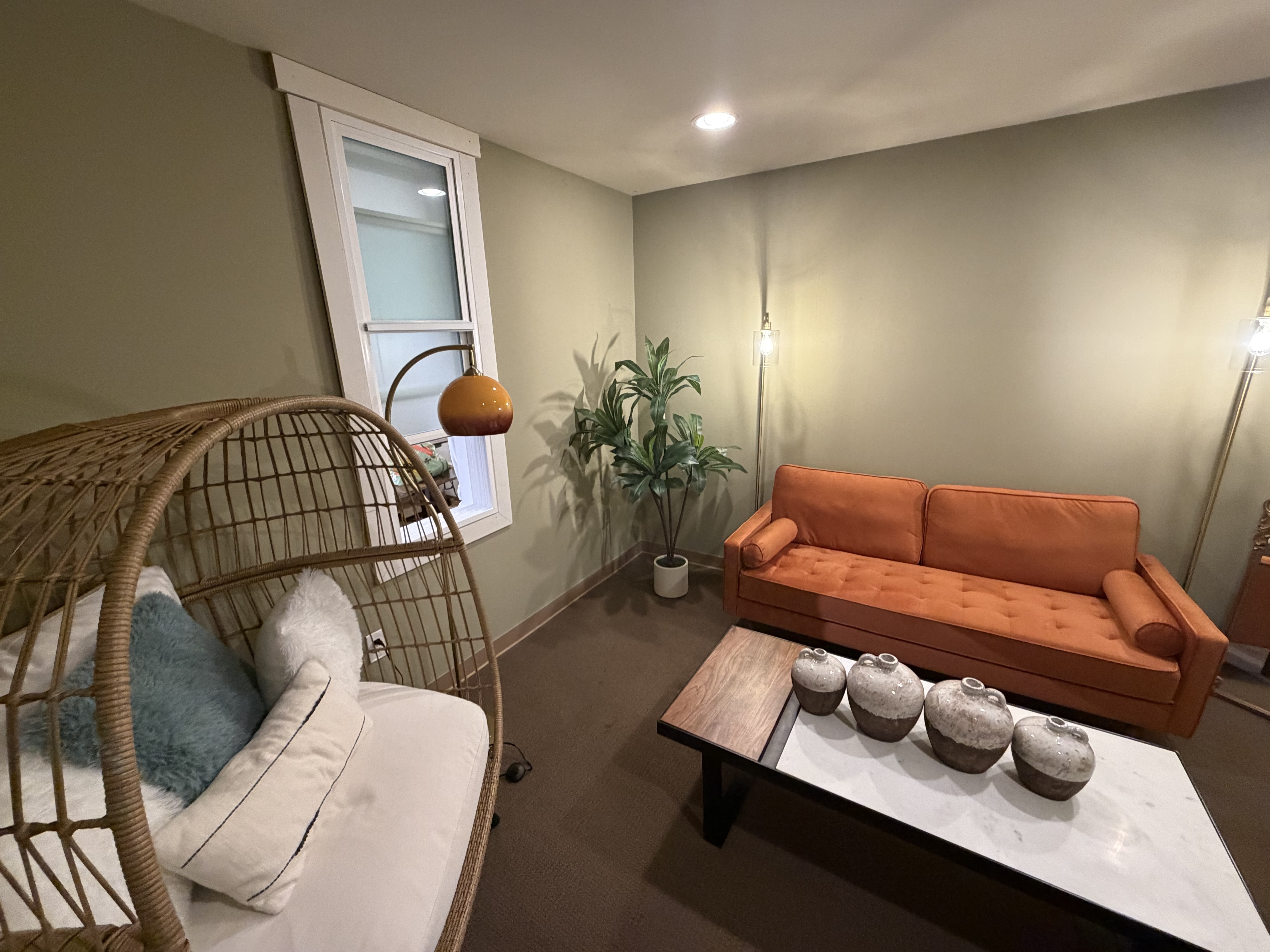 Wide view of the Mister Postman studio lounge with orange sofa, hanging wicker chair, coffee table, plants, and floor lamps